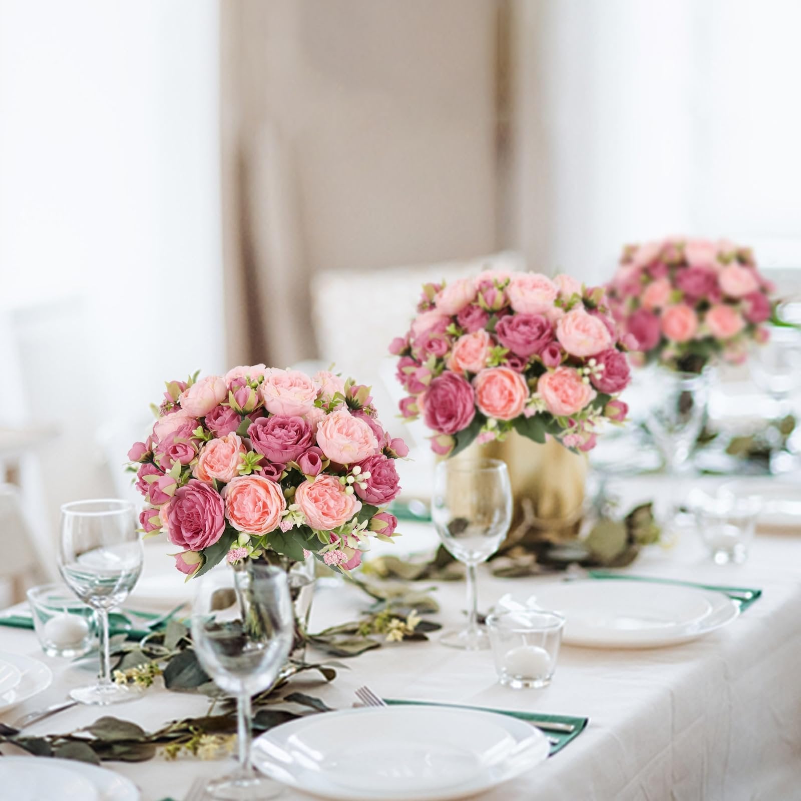 Table setting with pink artificial peony bouquets in gold and silver vases, plates, glasses, and candles.