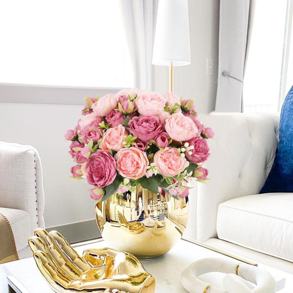 Pink artificial peony bouquet in a gold vase on a table with decorative items.