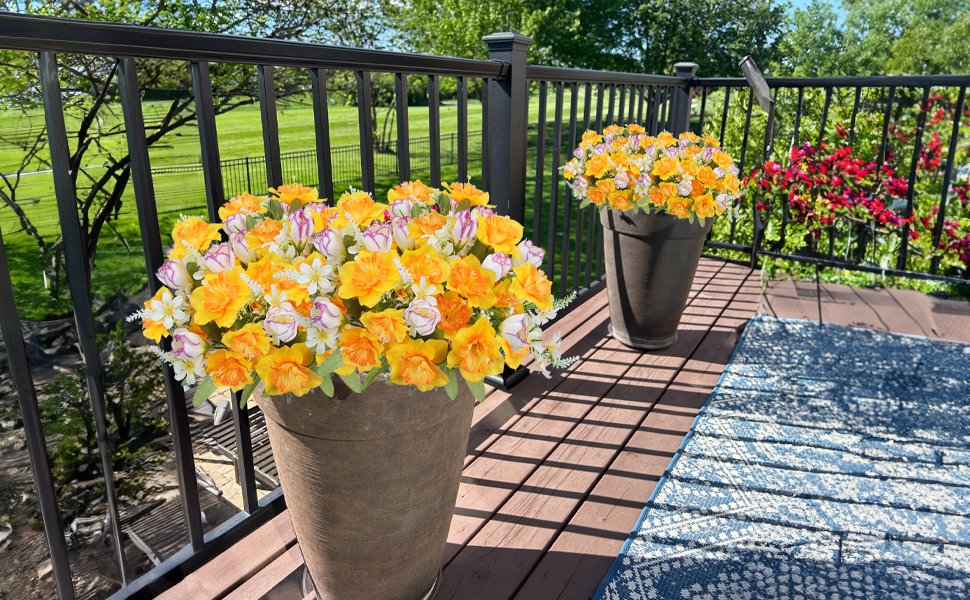 Two large planters filled with artificial daffodils and tulips on a wooden deck with a railing and a blue rug.