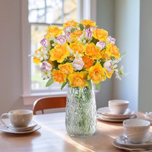 Artificial daffodils and tulips in a glass vase on a wooden table with place settings.