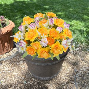 Artificial daffodils and tulips in a dark gray planter pot, set outdoors on mulch with green grass in the background.
