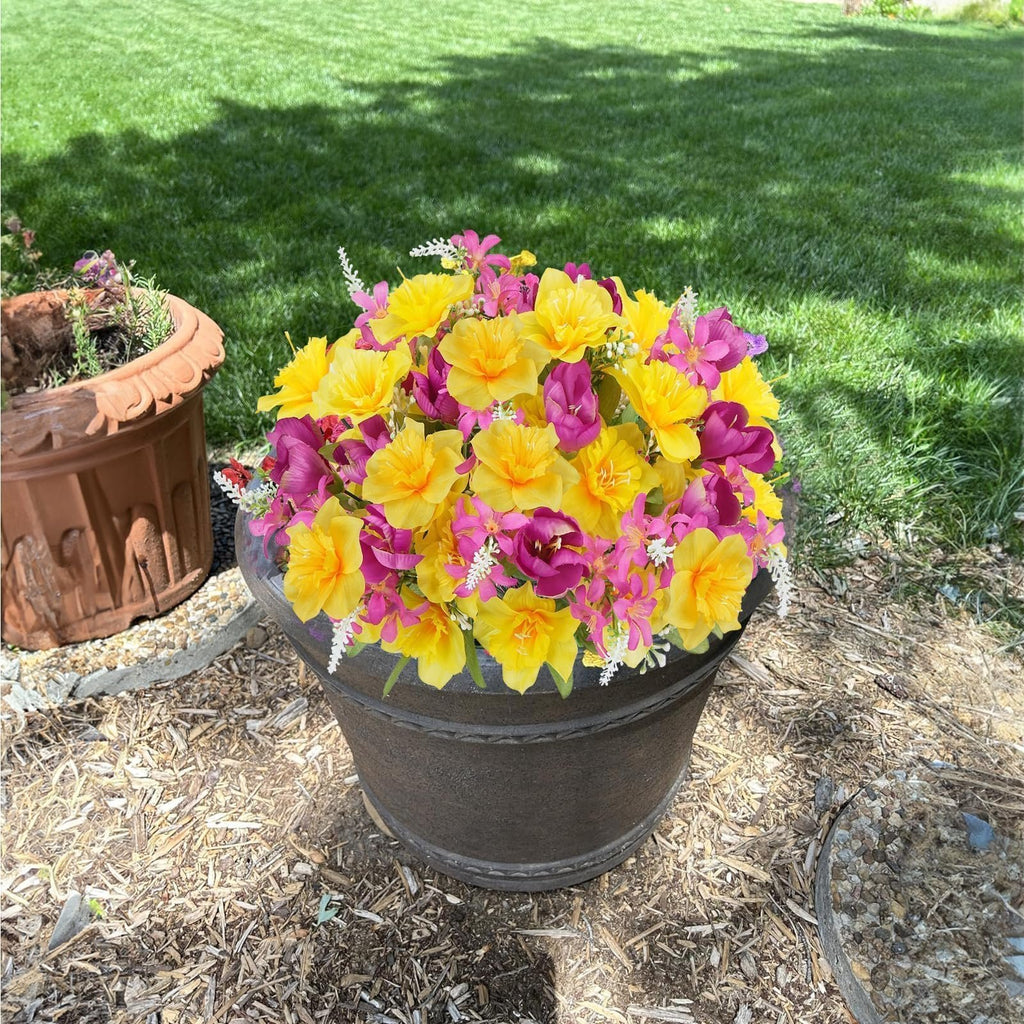 Bouquet of yellow daffodils and pink tulips in a dark pot on mulch with grass in the background