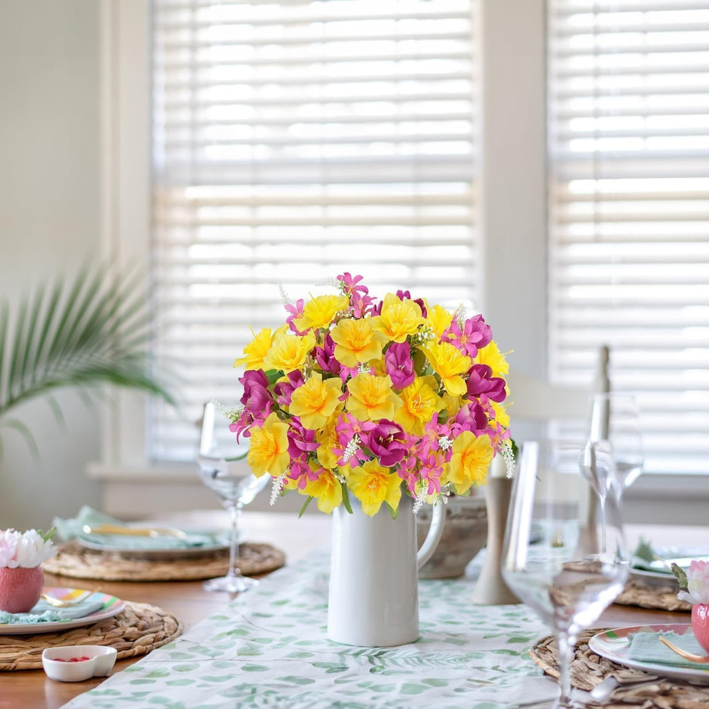 Bouquet of yellow daffodils and pink tulips in a white vase on a table runner with a leaf pattern.