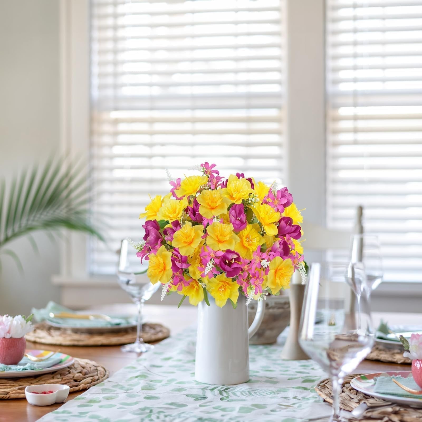 Bouquet of yellow daffodils and pink tulips in a white vase on a table runner with a leaf pattern.