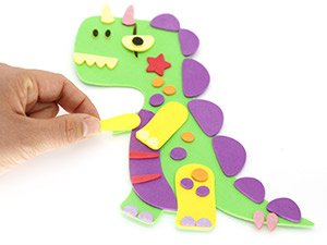 Hand attaching a yellow foam arm to a green foam dinosaur craft kit on a white background.