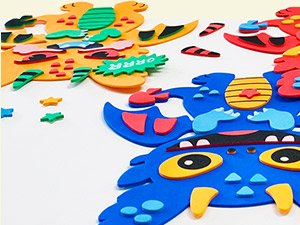 Close-up of colorful foam art craft kit pieces, including monster shapes and small decorative foam stickers, on a white background.