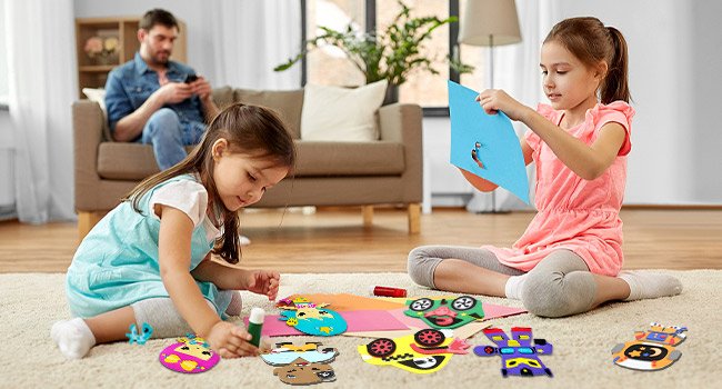 Two young girls crafting foam butterfly art projects on a rug, with a man in the background on a couch.