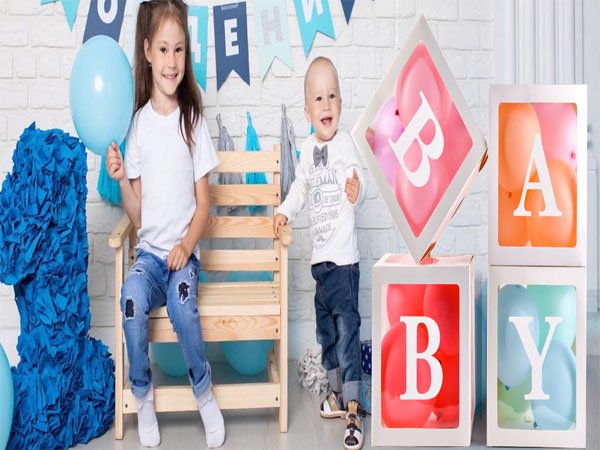 Two children posing with clear balloon boxes spelling BABY for a party decoration. A large blue number 1 is on the left.