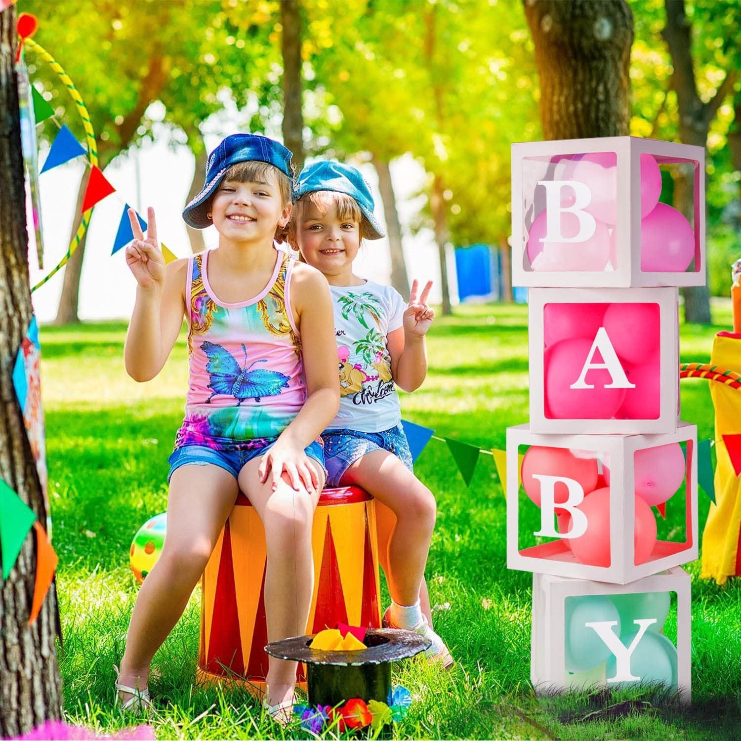 Two smiling girls in hats sit on a drum next to stacked clear boxes spelling BABY with balloons inside.