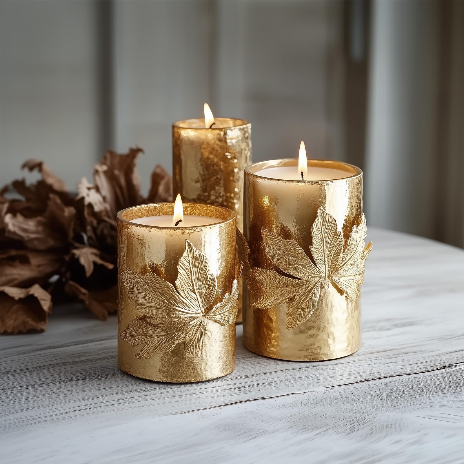 Three lit gold pillar candles with embossed leaf designs on a rustic wooden table with dried leaves