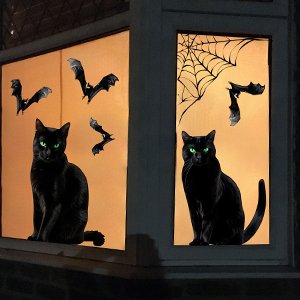 Two black cats with green eyes sit on windowsills with Halloween window clings of bats and a spiderweb.