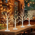 Four lighted white birch tabletop trees with warm white LEDs arranged on a wooden table, with a blurred background of string lights and a building.