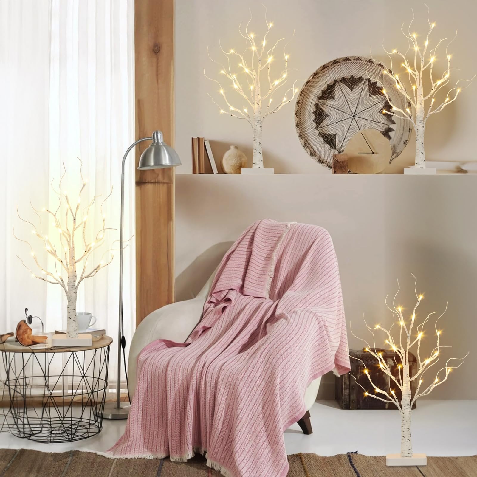 Four lighted birch tabletop trees with warm white LEDs arranged in a cozy living room setting with a chair and blanket.