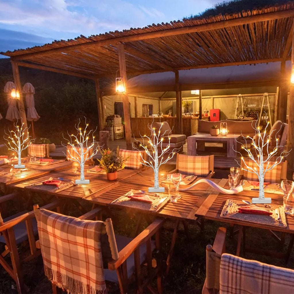 Four lighted birch tabletop trees with warm white LEDs set on a wooden table outdoors at dusk.