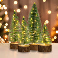 Four mini artificial Christmas trees with warm white LED lights and wooden bases on a table with bokeh background.