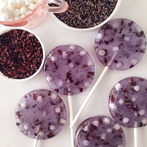 Six clear lavender lollipops with dried flower pieces and white cubes, arranged around bowls of dried lavender, cocoa nibs, and mini marshmallows.