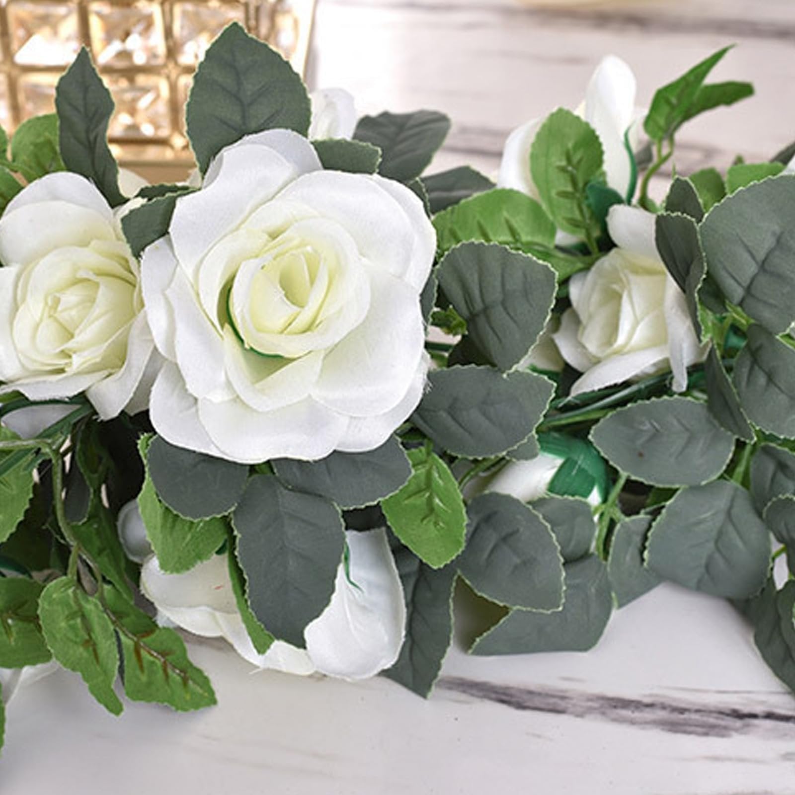 Artificial white rose garland with green leaves draped on a marble surface, close-up view.