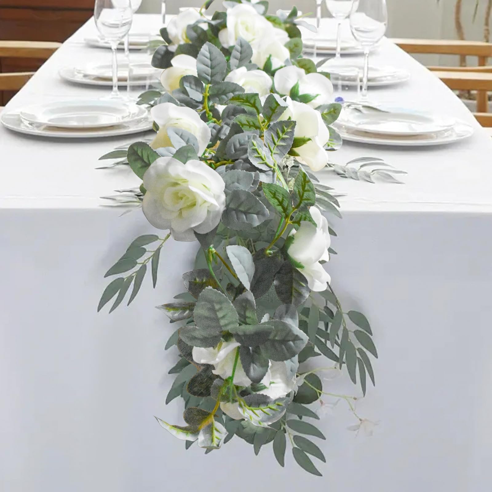 Artificial rose garland with white flowers and green leaves draped across a white tablecloth on a dining table.