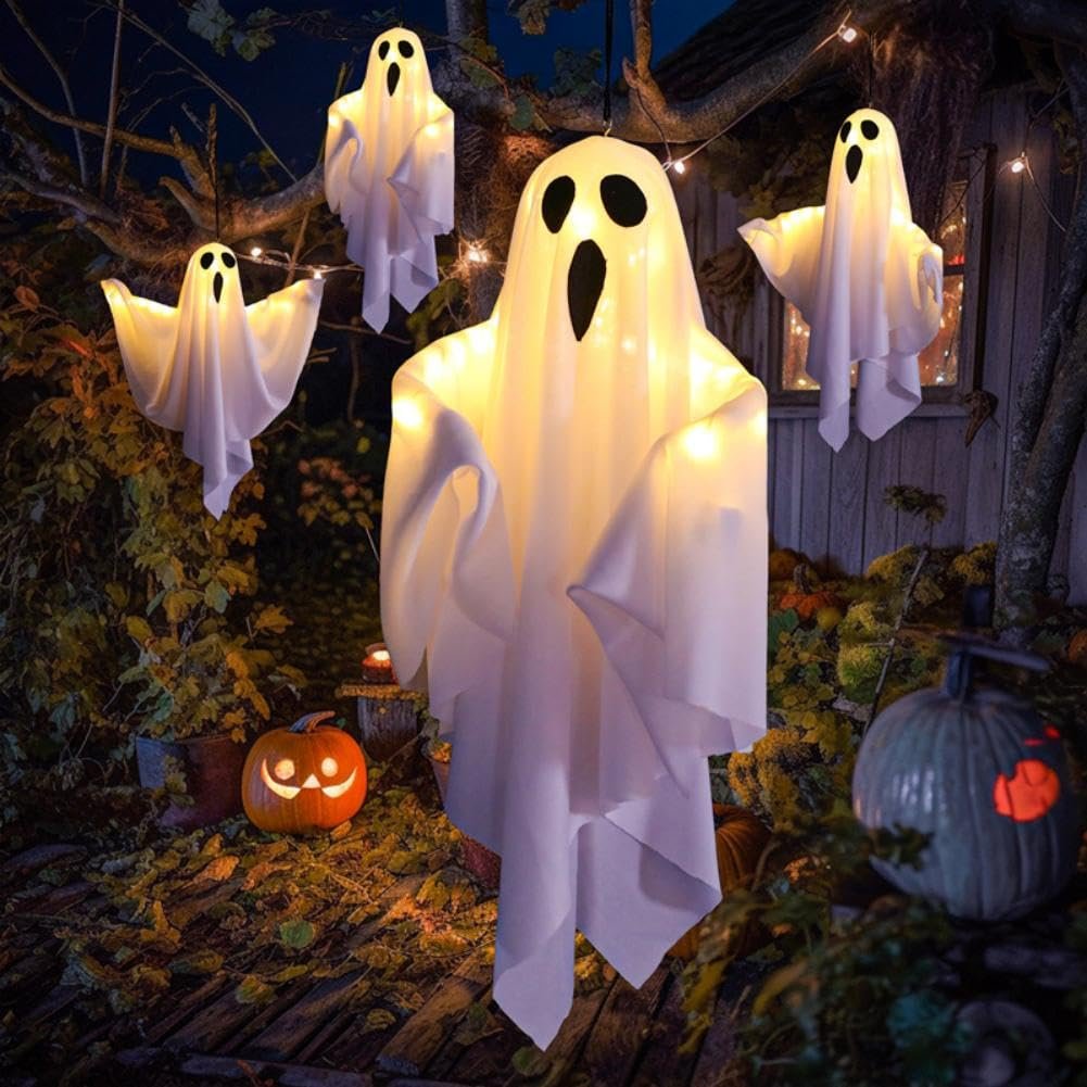 Four lighted white fabric ghost decorations hanging outdoors at night with pumpkins and string lights