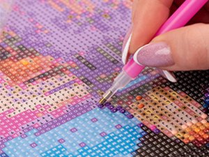 Close-up of a hand using a pink tool to place a diamond onto a 5D diamond painting kit with a floral pattern.
