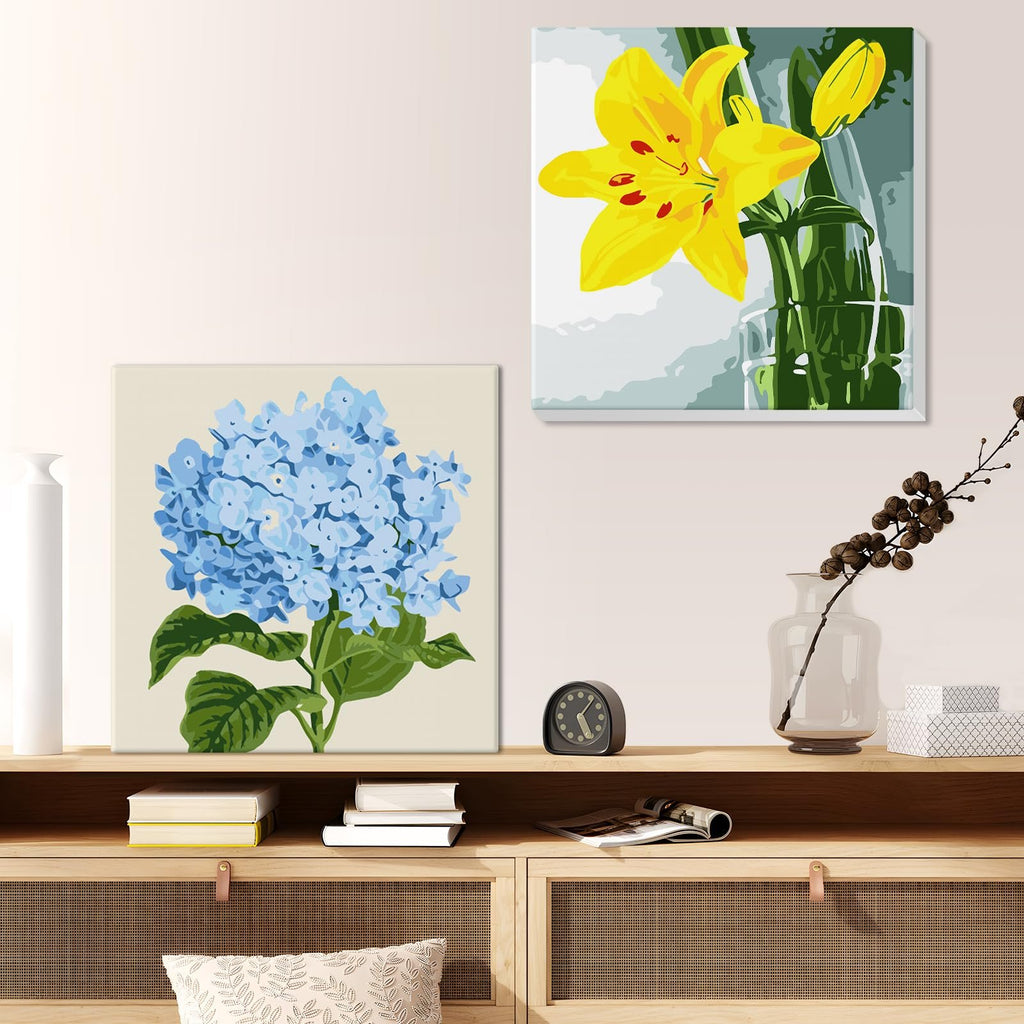 Two framed paint by number canvases, one with blue hydrangeas and one with a yellow lily, displayed on a shelf.