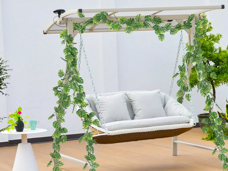Artificial monstera vines draped over a white swing set on a patio, with a white table and plant to the left.