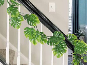 Artificial monstera vines draped along a staircase railing with white balusters and carpeted steps.