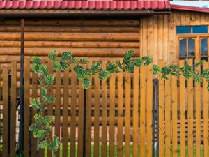 Artificial monstera vine with green leaves draped over a wooden fence in front of a log cabin.