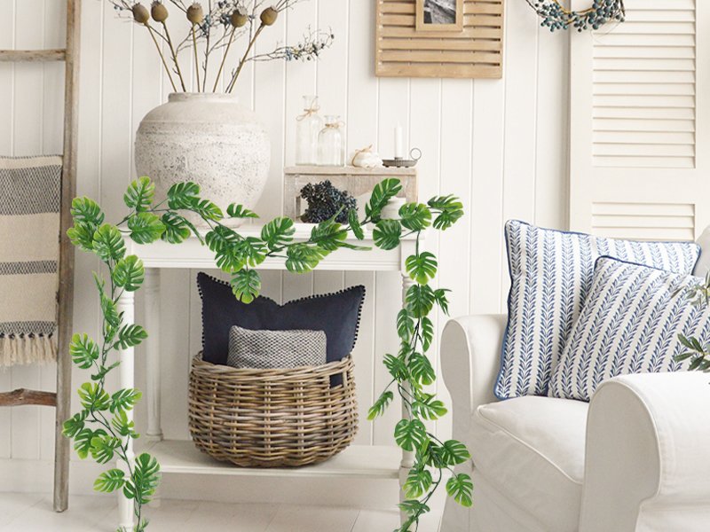Artificial monstera vines draped over a white table with decorative items and pillows on a sofa.