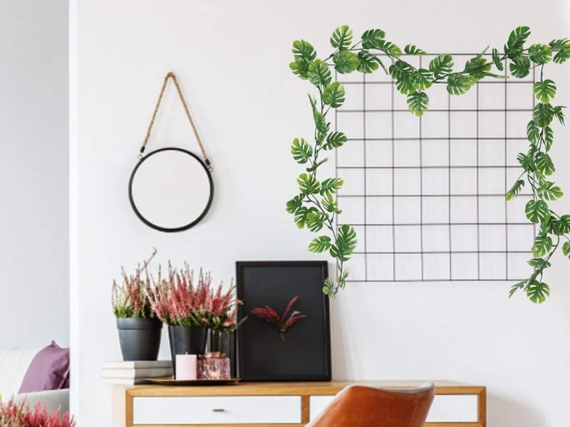 Artificial monstera vines draped over a grid on a white wall above a desk with plants and a mirror.