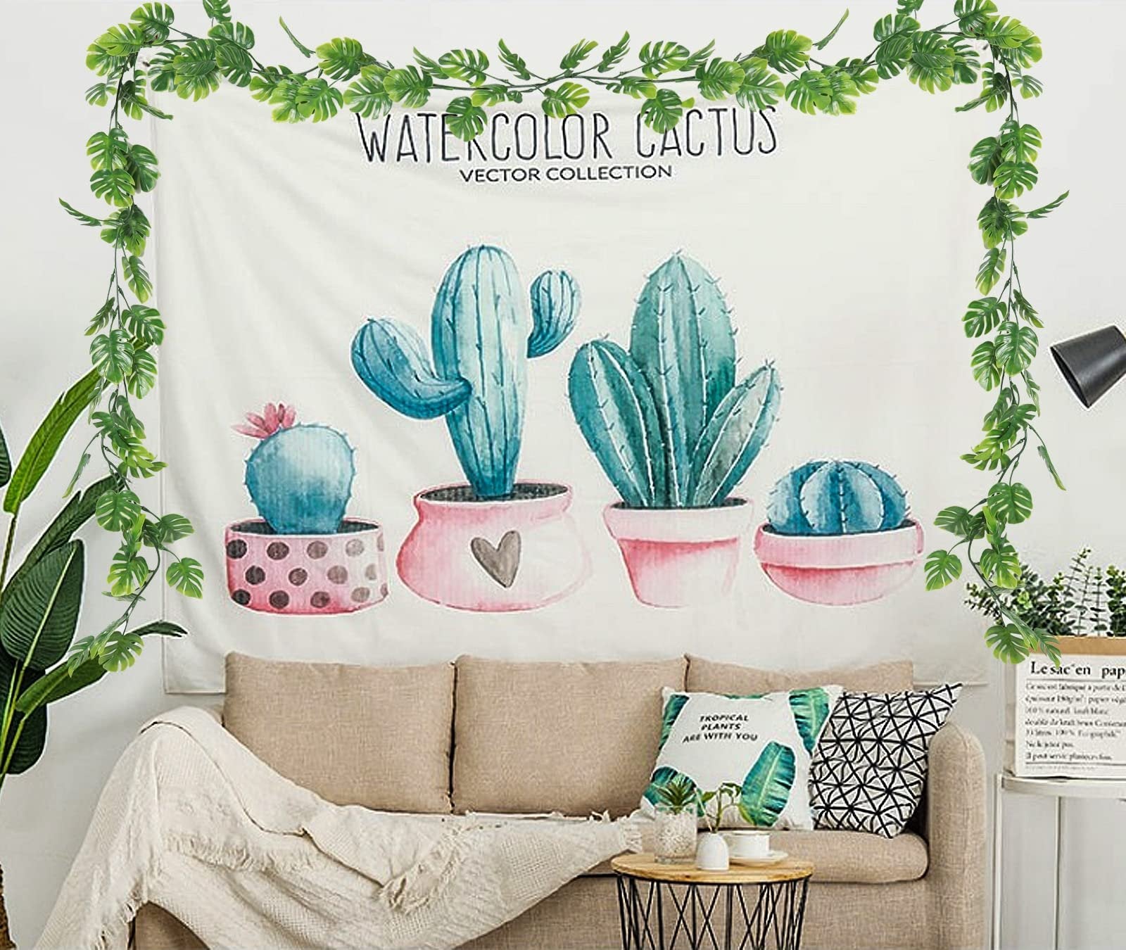 Artificial monstera vines draped around a tapestry featuring watercolor cacti in pots above a sofa.