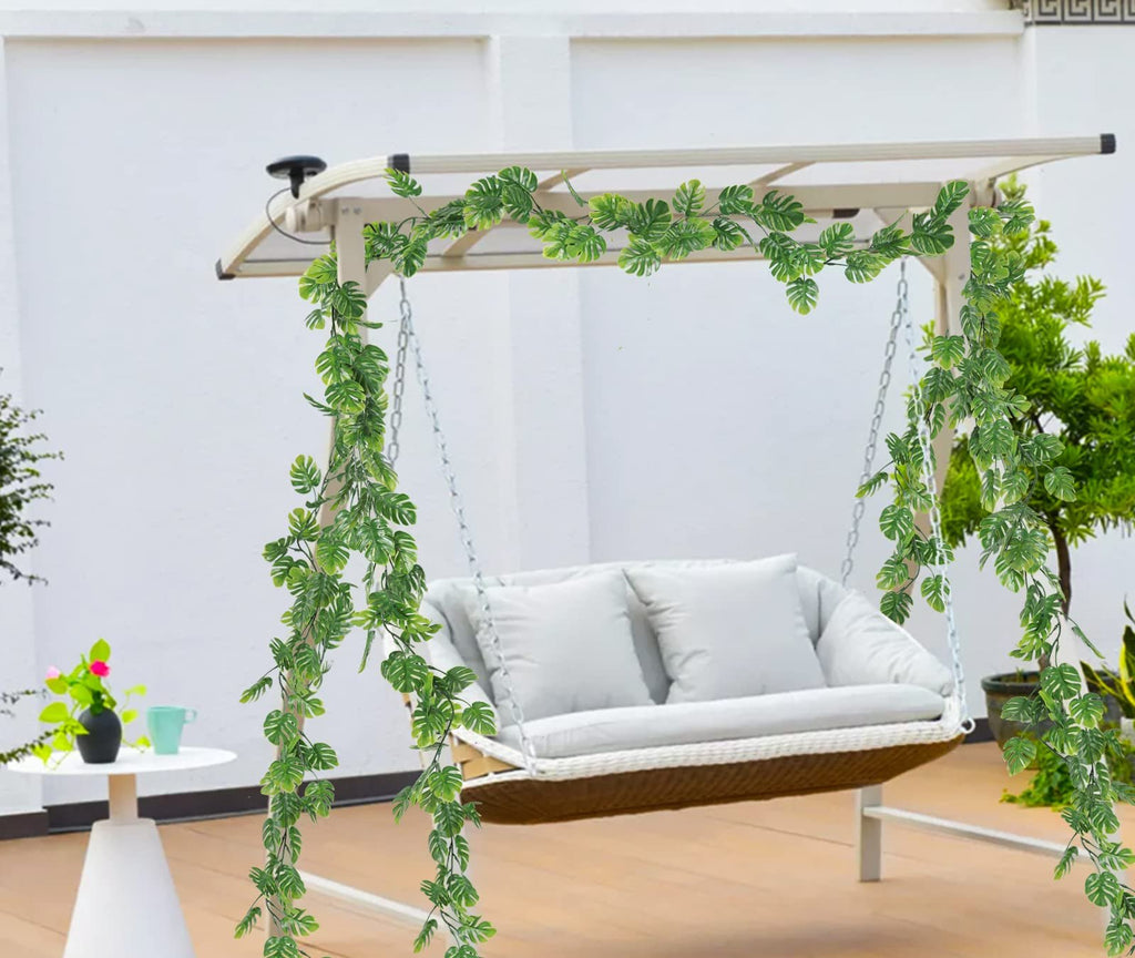 Artificial monstera vines draped over a white swing set with a cushioned seat, outdoors.
