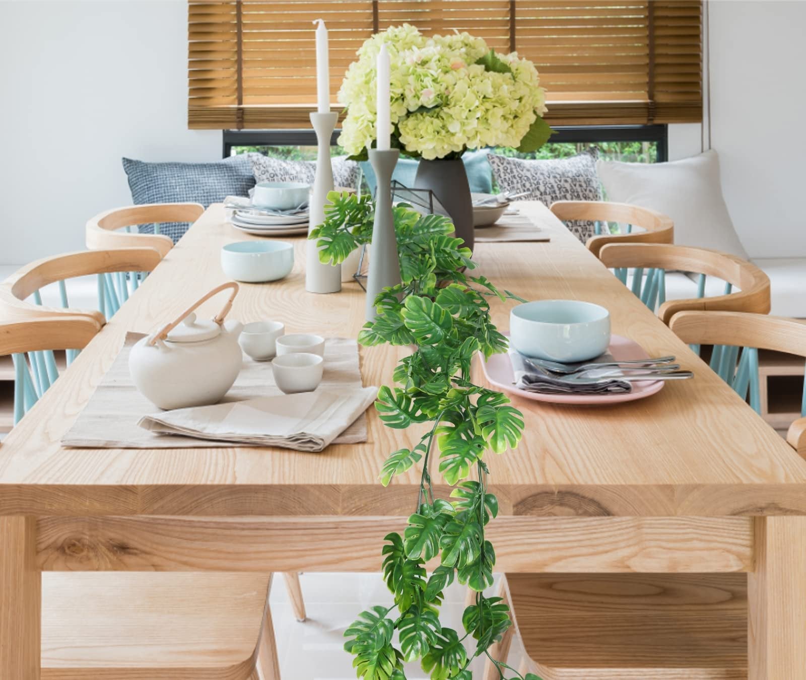Artificial monstera vines draped over a wooden dining table set for a meal, with place settings, a teapot, and floral centerpiece.