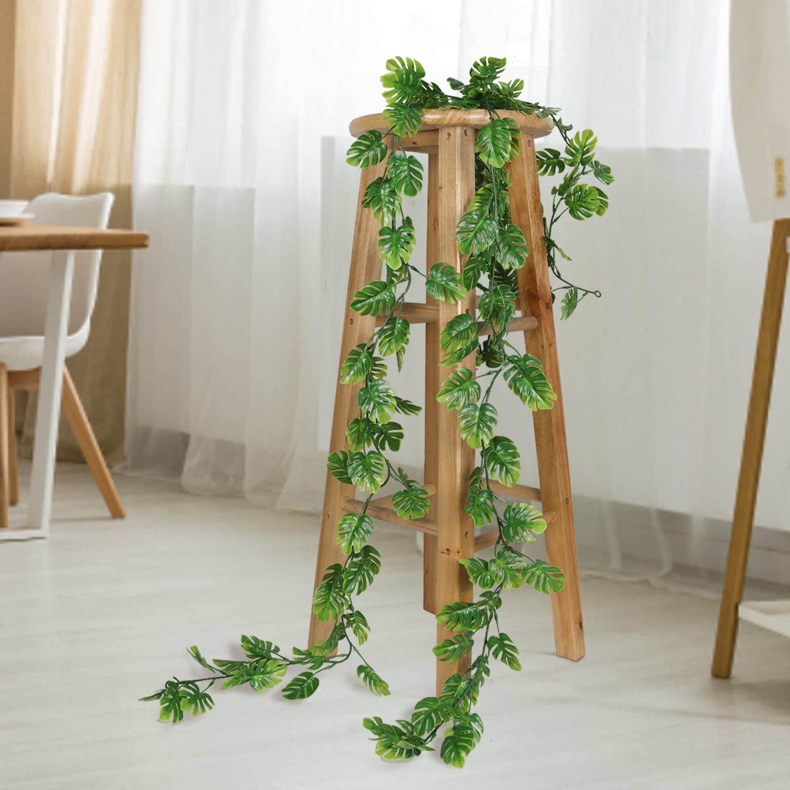 Artificial monstera vines draped over a wooden stool in a bright room with sheer curtains.