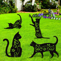 Four black metal cat silhouette garden stakes with vine and flower cutouts on green grass, with Halloween decorations in background.