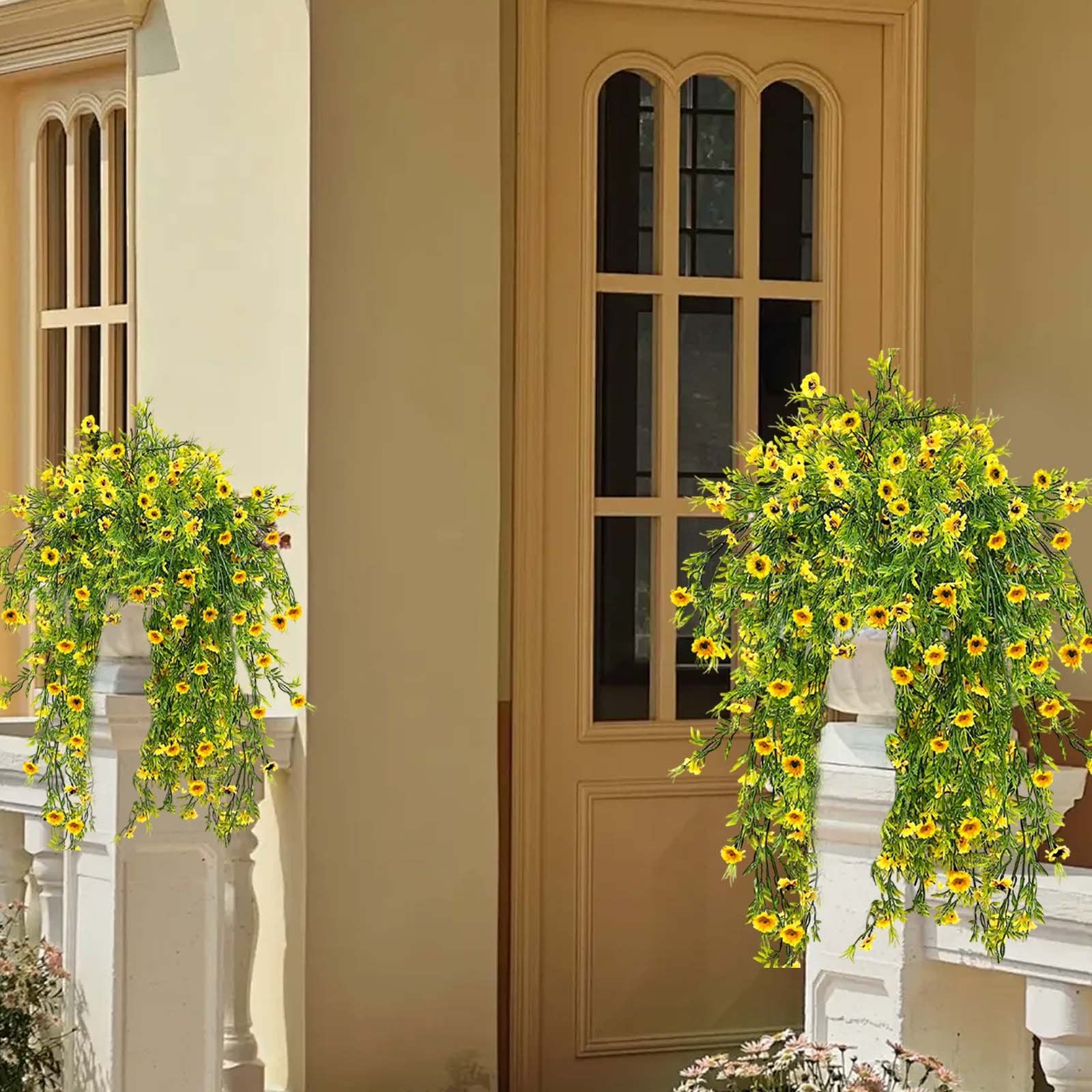 Two hanging baskets of bright yellow artificial daisy plants with green foliage, displayed on a porch railing next to a door.