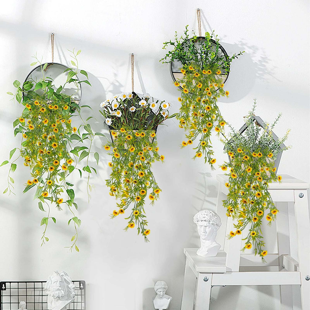 Four hanging artificial daisy plants with yellow flowers and green leaves displayed on a white wall.