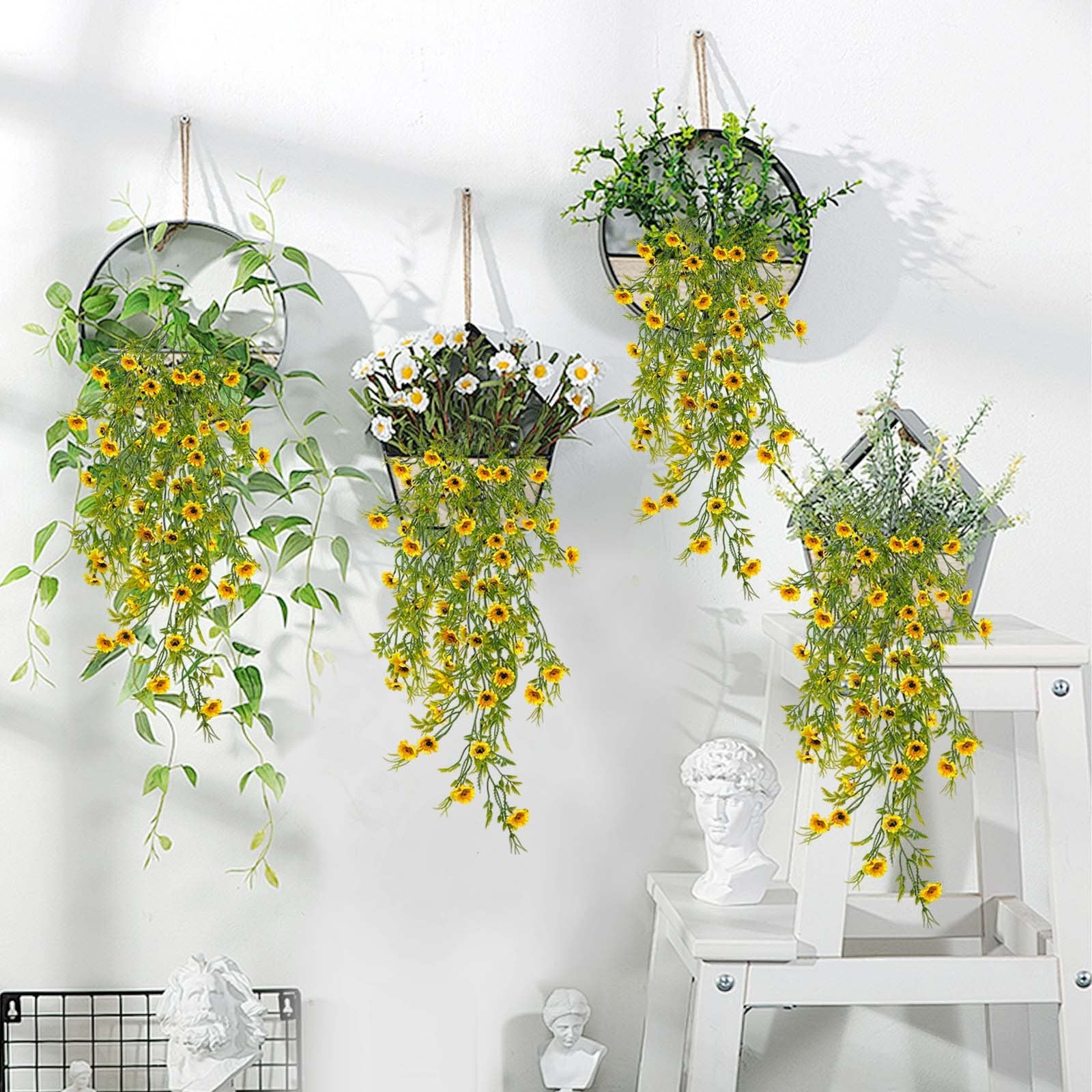 Four hanging artificial daisy plants with yellow flowers and green leaves displayed on a white wall.