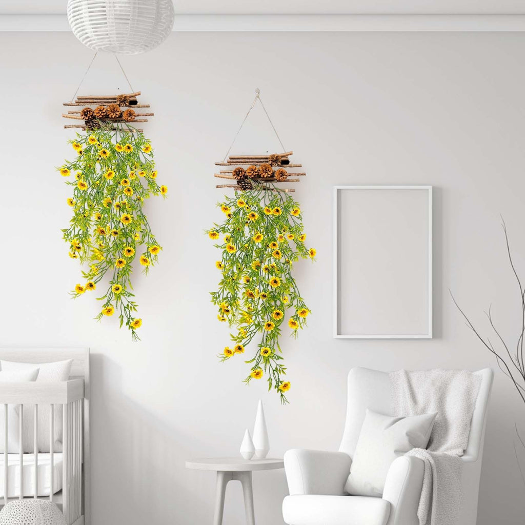 Two hanging artificial daisy plants with yellow flowers and pinecones on a white wall, next to a crib and armchair.