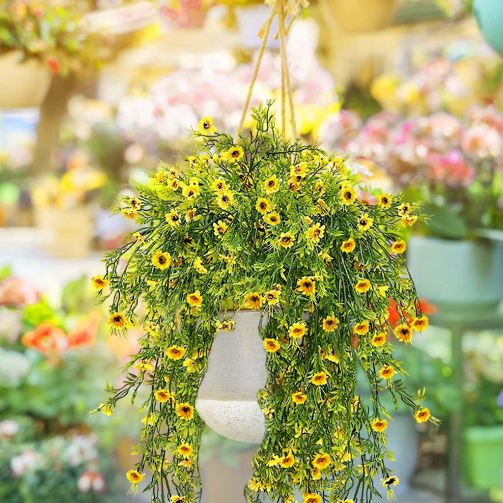 Hanging artificial daisy plant with bright yellow flowers and green foliage in a light gray pot, suspended by rope.
