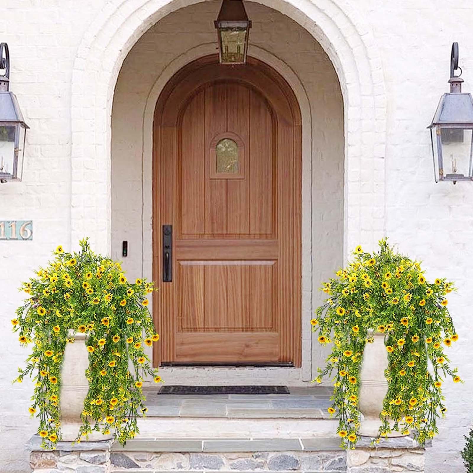 Two hanging baskets filled with bright yellow artificial daisy plants cascading down stone planters on a porch.