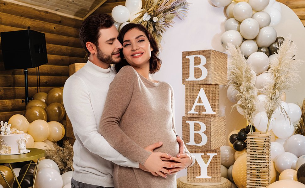Pregnant couple embracing near stacked burlap blocks spelling BABY, decorated for a baby shower with balloons and a cake.