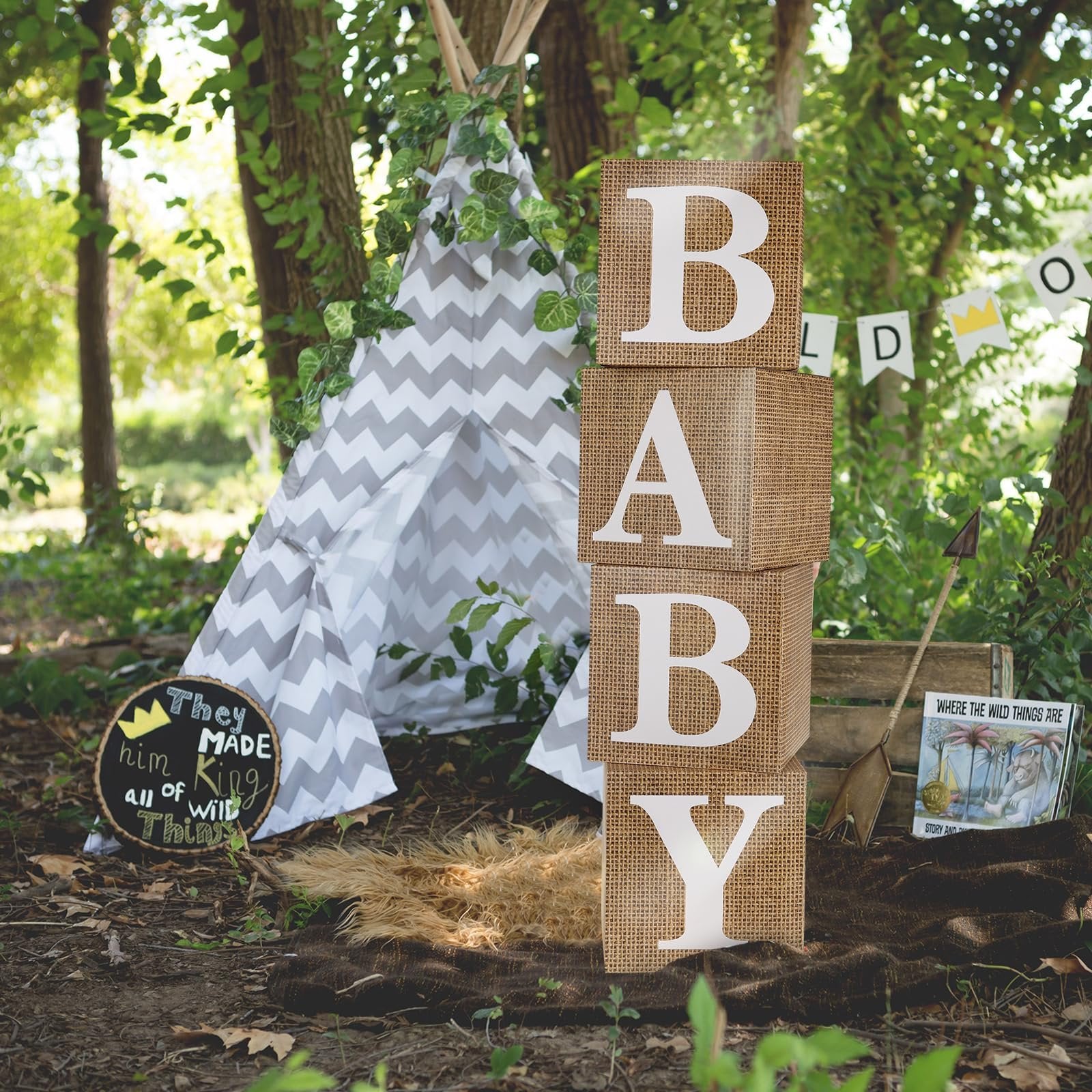 Four burlap baby blocks spelling 'BABY' stacked in a forest setting with a teepee and decorations.