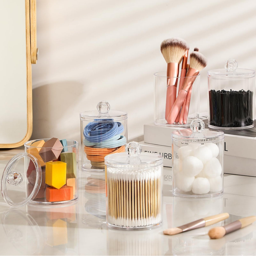 Set of clear acrylic canisters filled with makeup sponges, hair ties, cotton swabs, cotton balls, and makeup brushes on a reflective surface.