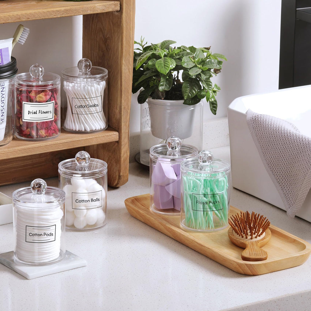 Four clear acrylic bathroom canisters with lids, filled with cotton pads, cotton balls, cotton swabs, and floss picks, arranged on a countertop.