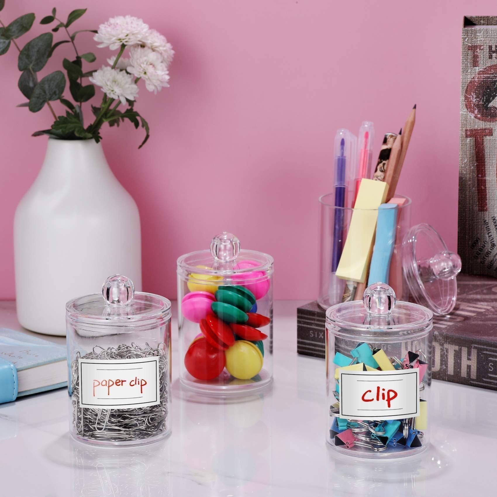 Three clear acrylic canisters filled with office supplies on a marble surface, with a vase of flowers and books in the background.