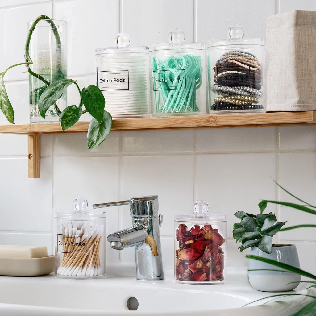 Four clear acrylic bathroom canisters on a shelf and sink, filled with cotton pads, floss picks, hair accessories, and rose petals.