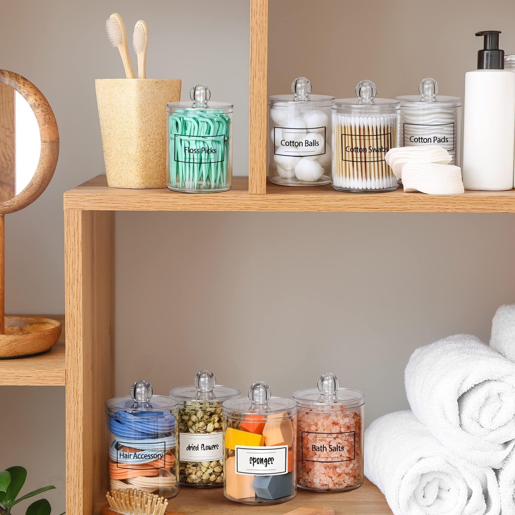Clear acrylic bathroom canisters filled with cotton balls, swabs, pads, floss picks, hair accessories, dried flowers, sponges, and bath salts on a wooden shelf.