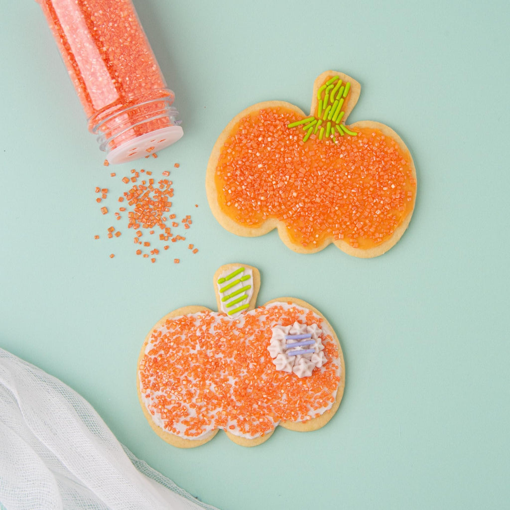 Two pumpkin-shaped sugar cookies decorated with orange sprinkles and frosting, with a clear sprinkle bottle spilling some sprinkles.
