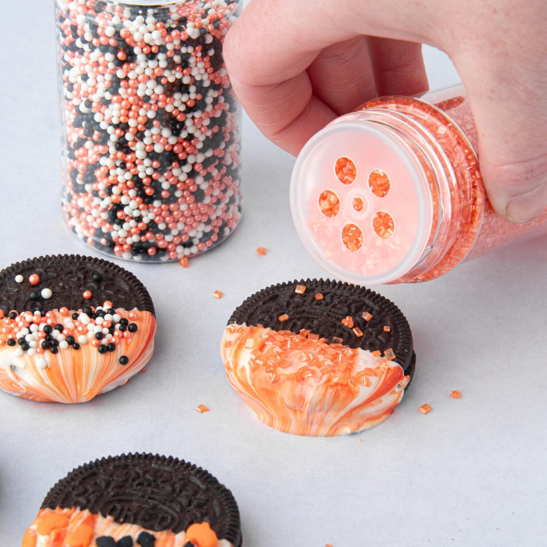 Hand pouring orange sprinkles onto a chocolate-dipped cookie with orange and white icing, next to a jar of Halloween-themed sprinkles.